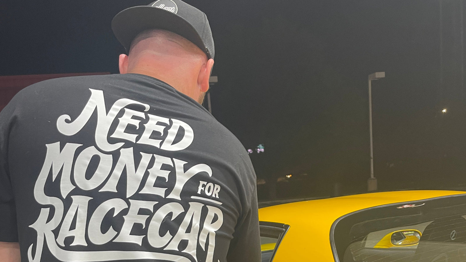 Person wearing a black t-shirt with 'Need Money for Racecar' text, standing next to a yellow car at night.