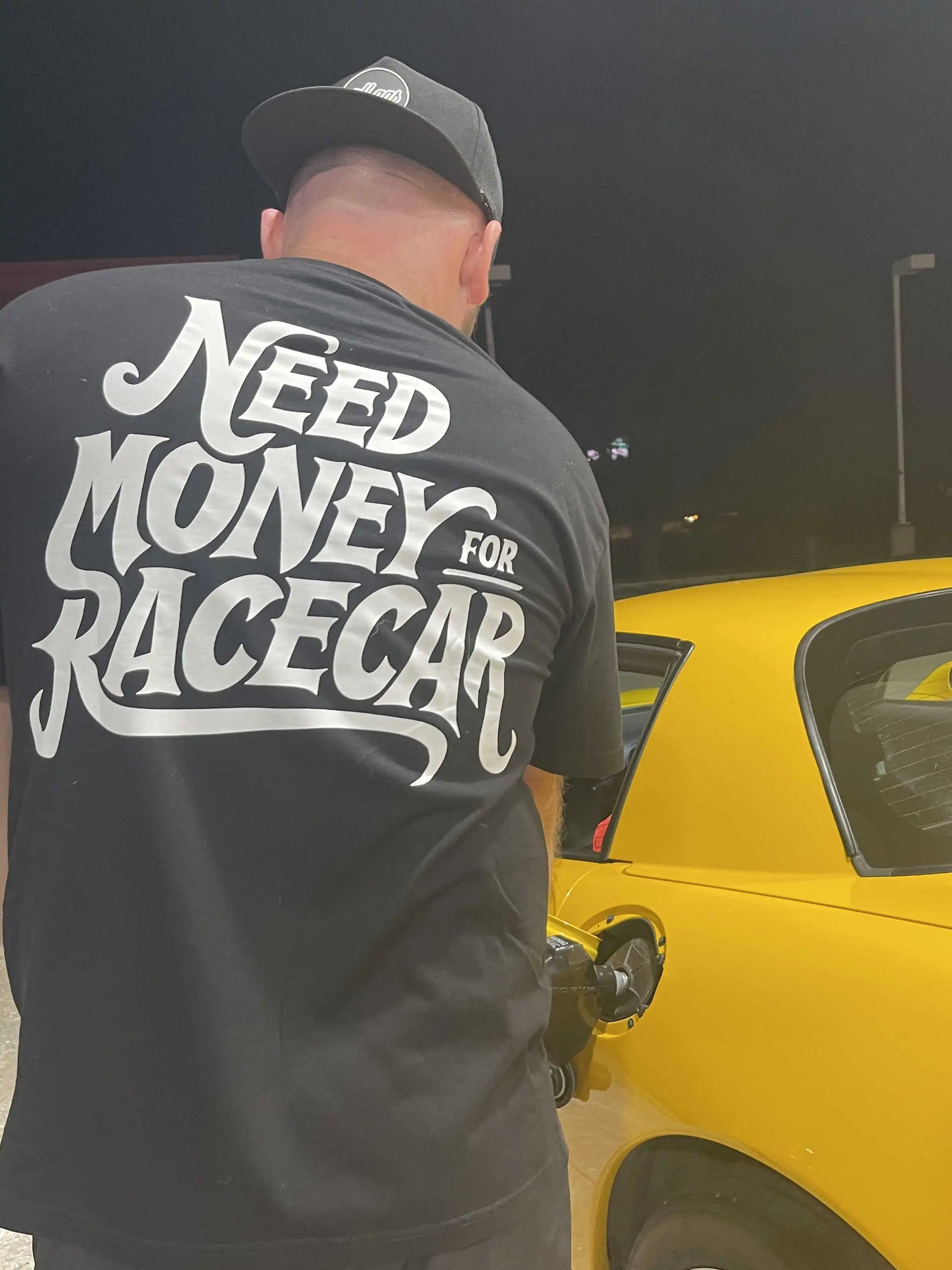 Person wearing a black t-shirt with 'Need Money for Race Car' text, fueling a yellow car at night.